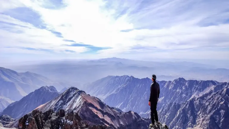 Para ayudarte con la imagen del artículo sobre el trekking al Toubkal, aquí tienes opciones de Alt Text, Title, Caption y Description pensadas para optimizarla: Opciones para la Imagen del Artículo Alt Text (Texto Alternativo) "Trekker ascendiendo el sendero rocoso del Monte Toubkal con picos nevados al fondo.