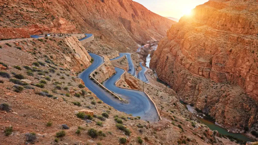 Pueblo bereber tradicional de adobe encaramado en una ladera rocosa del Atlas marroquí, con casas color tierra y palmeras verdes bajo un cielo azul.